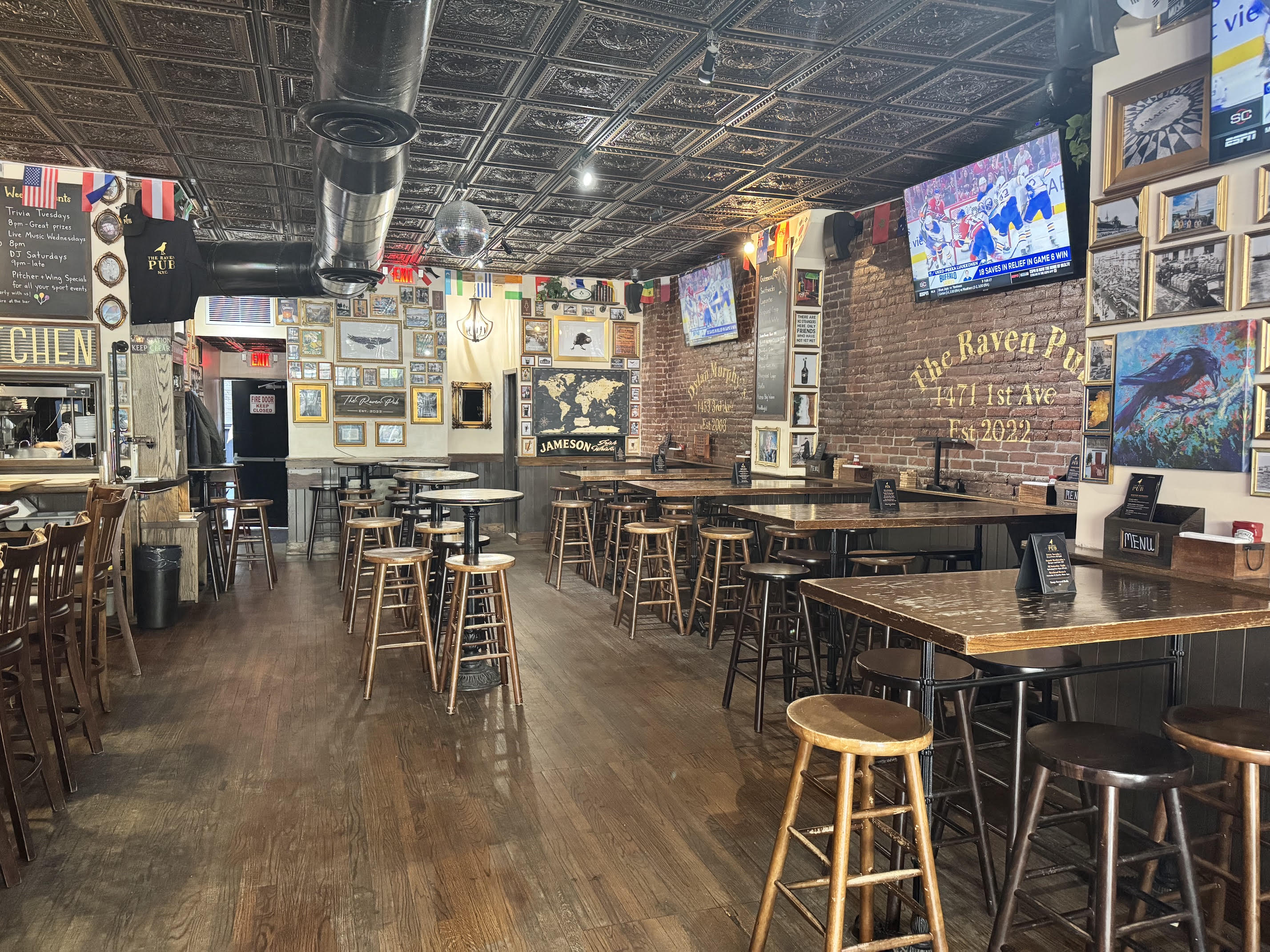 A cozy pub with wooden tables and stools, brick walls covered in framed photos and decor, a tin ceiling, two TVs, and international flags hanging above. The space is empty and well-lit with natural light.