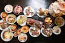 An overhead view of a dark table filled with assorted dishes, including steaks, seafood, salads, sauces, breads, and various sides, displayed on white plates and bowls for a large, diverse meal.