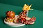 A gourmet hamburger with lettuce, tomato, pickles, and melted cheese on a seeded bun, paired with a whole cooked lobster and a side of French fries in a metal cup, set against a green background.