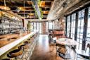 A rustic bar interior with a long marble counter lined with stools, exposed brick walls, and wooden ceiling beams. The space features leather booth seating on the right and large windows letting in natural light.