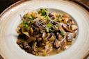 A plate of creamy pasta topped with sautéed mushrooms and microgreens, served in a brown-rimmed dish. The sauce appears rich, and the dish is garnished with fresh herbs.