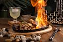 A sizzling platter of stir-fried meat and vegetables is set on a wooden table, with flames rising above the dish. A glass of a light-colored beverage and chopsticks are nearby, and decorative stones surround the plate.