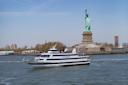 A white City Cruises ferry boat sails past the Statue of Liberty on a sunny day, with passengers on deck and the New York City skyline in the background.