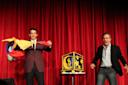 Two men perform a magic act on stage in front of closed red curtains. One man holds colorful scarves, and the other stands beside a decorated box on a small table. Both are dressed in suits and look engaged in the performance.
