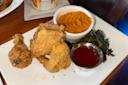 A plate with fried chicken pieces, a small bowl of mashed sweet potatoes, cooked greens, and a metal cup of red dipping sauce, all served on a white rectangular plate on a wooden table.
