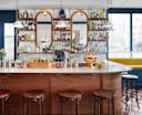 A stylish bar with a polished wooden counter, four round stools, glassware, and bottles of liquor neatly arranged on shelves in front of three mirrors. Large windows provide natural light, and pendant lights hang above the bar.