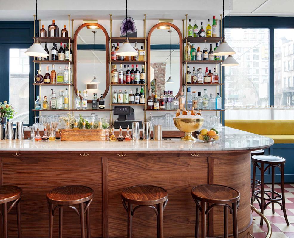 A stylish bar with a polished wooden counter, four round stools, glassware, and bottles of liquor neatly arranged on shelves in front of three mirrors. Large windows provide natural light, and pendant lights hang above the bar.