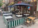 Outdoor seating area of a restaurant with tables and chairs, some set for dining. Two people sit at one table. Green and white barriers enclose the area. Building storefronts and sidewalk visible in the background.