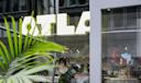 A man wearing glasses sits inside a modern café, partially obscured by a green plant. Large, bright letters spelling "OTLA" are visible on the glass window, with city reflections outside.