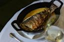 A whole baked fish in a black oval dish with creamy sauce and herbs, served on a white tablecloth next to a glass of white wine and a silver spoon.