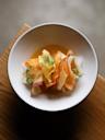 A white bowl contains thin ribbons of colorful root vegetables, like carrots and radishes, garnished with fresh herb sprigs and set on a wooden surface.