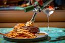 A hand uses a siphon to top a steak with foam, next to a serving of fries, dipping sauce, and a seeded bun on a plate; a martini with olives is in the background on a teal table.