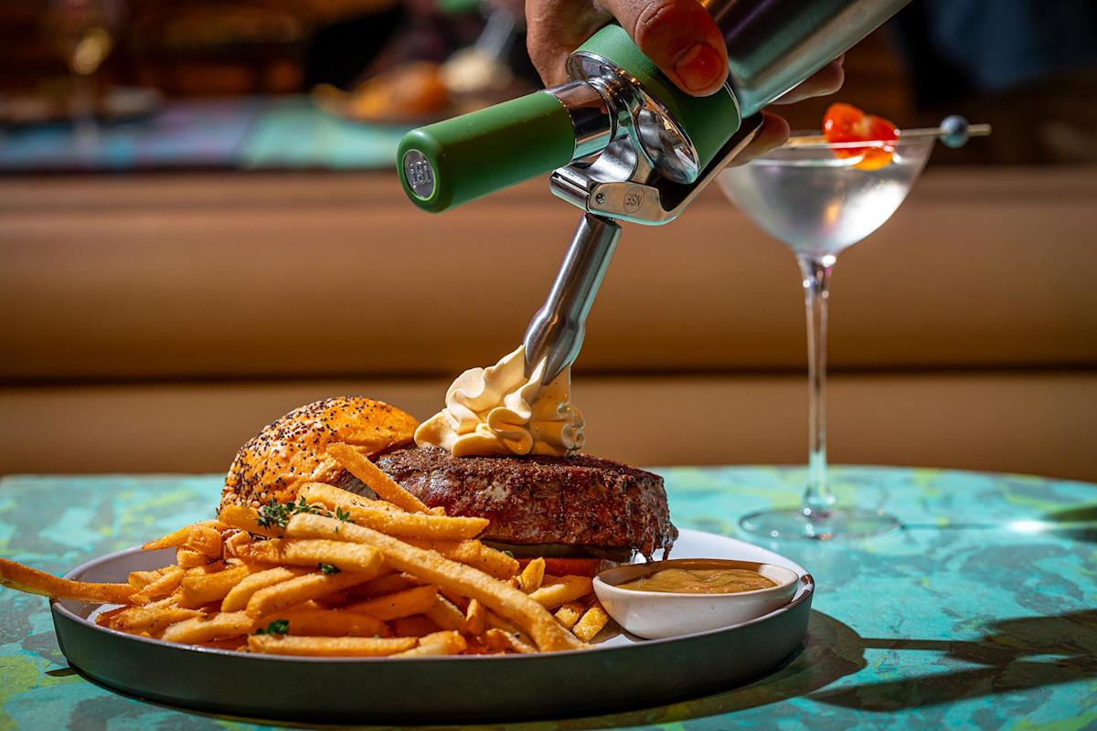 A hand uses a siphon to top a steak with foam, next to a serving of fries, dipping sauce, and a seeded bun on a plate; a martini with olives is in the background on a teal table.