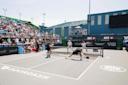 Four athletes play a competitive pickleball doubles match on an outdoor court, with a large crowd of spectators watching from bleachers under a sunny sky. Sponsor logos are visible around the court.