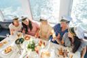 Five women wearing captain hats sit around a table on a boat, laughing and enjoying food and drinks with a view of sparkling water through the windows behind them.