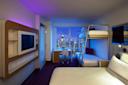 A modern hotel room with a large bed, bunk beds, a wall-mounted TV, a desk, and a city skyline view through large windows at night, illuminated by purple and blue accent lights.