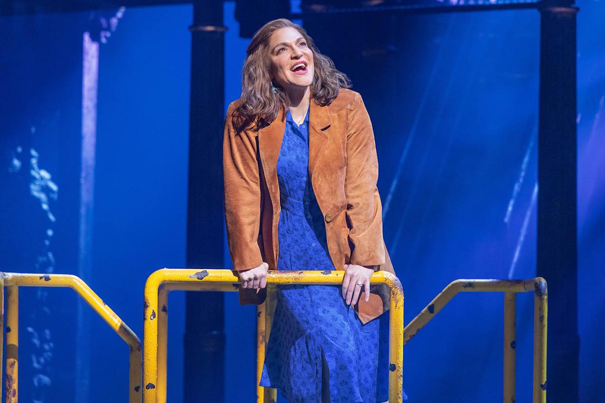 A woman in a blue dress and brown jacket stands behind a yellow railing on stage, with blue and purple lighting creating a dramatic atmosphere.