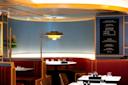A modern diner interior with red booth seating, set tables, and a blackboard menu on the wall. The space features a unique ceiling design and a hanging light fixture. A plant decorates the corner, adding a touch of greenery.