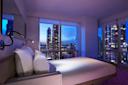 A modern hotel room with large windows offers a night view of a city skyline with tall buildings and lights. The room features a neatly made bed with white linens and a reading lamp.