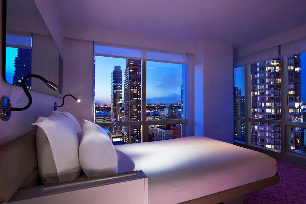 A modern hotel room with large windows offers a night view of a city skyline with tall buildings and lights. The room features a neatly made bed with white linens and a reading lamp.