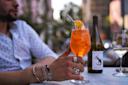 A person wearing a polka dot shirt and silver rings holds a glass of an orange cocktail with a slice of orange and a straw on a table outside. There are wine bottles and glasses in the background.