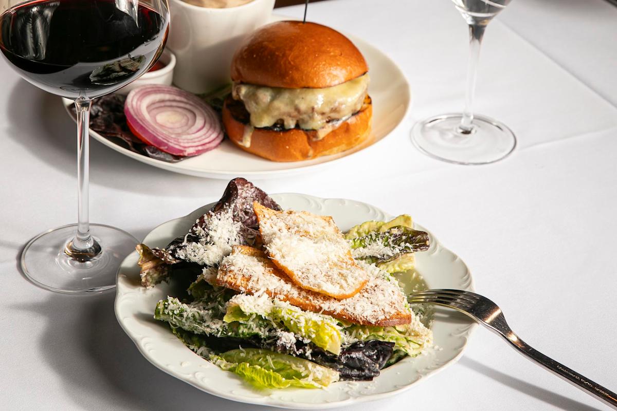 A plate of Caesar salad with grilled chicken, a fork, a glass of red wine, and a cheeseburger with onions and pickles in the background on a white tablecloth.