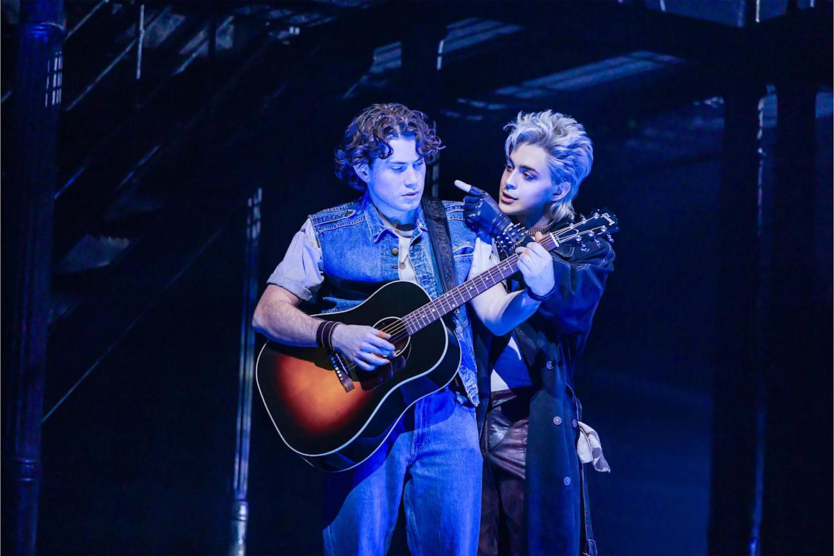 Two performers on stage: one plays an acoustic guitar while the other, standing close behind, gestures near the strings. Both are under dramatic blue lighting, with stairs and structural beams in the background.