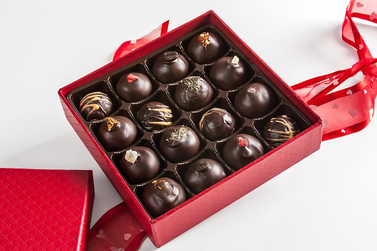 A red gift box filled with assorted chocolate truffles, each decorated with different toppings, sits open on a white surface. A red ribbon with heart patterns is attached to the box.