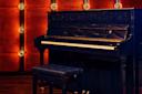 A dark blue upright piano with a matching padded bench sits on a wooden floor. The background features a warm, orange-brown wall with circular lights at regular intervals, creating a cozy ambiance.