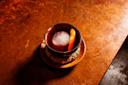 A cocktail with a large round ice cube and an orange peel garnish sits in a rustic cup on a coaster and plate, atop a textured brown surface.