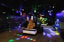 A dimly lit nightclub with colorful disco lights, black walls, bar tables, and a large teddy bear sitting on a couch in the center of the room. Green potted plants add decoration.
