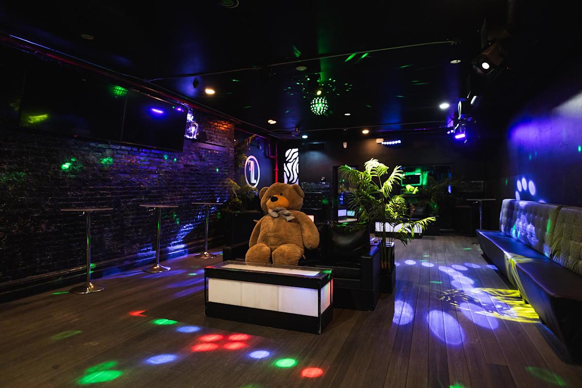 A dimly lit nightclub with colorful disco lights, black walls, bar tables, and a large teddy bear sitting on a couch in the center of the room. Green potted plants add decoration.