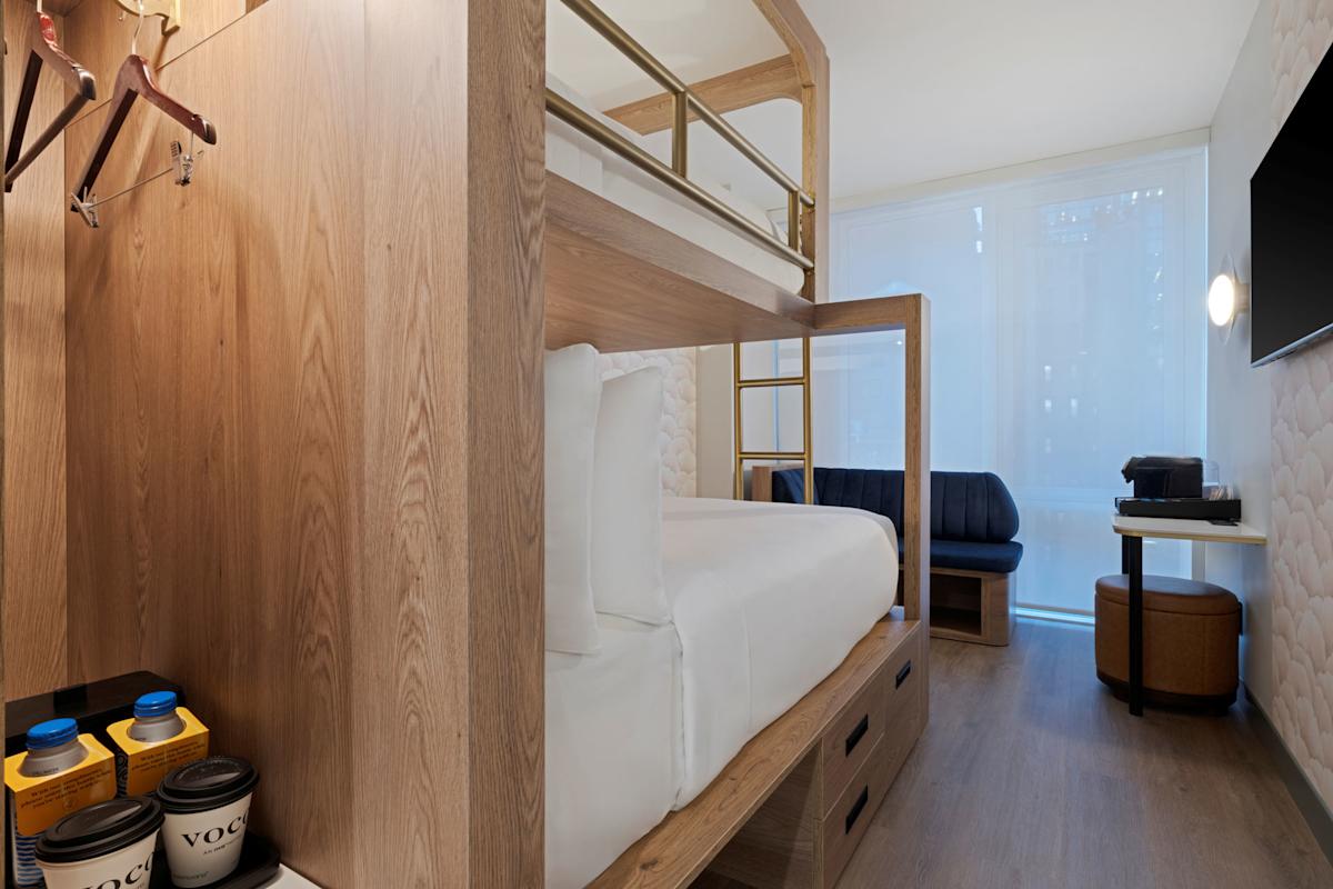 Modern hotel room with a wooden bunk bed, wall-mounted TV, coffee maker, small sofa, and floor-to-ceiling window with sheer curtains. Hanging space with hangers and cups is visible on the left side.