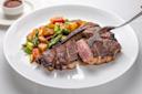 A grilled steak, sliced to show a pink center, is served with roasted mixed vegetables including asparagus, potatoes, and tomatoes on a white plate. A fork is holding a piece of steak; a small bowl of sauce is on the side.