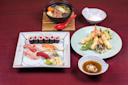 A Japanese meal with assorted sushi and sashimi, tempura vegetables and shrimp, a bowl of soup with vegetables and tofu, dipping sauce, and a small bowl of dipping condiment, all on a red table.