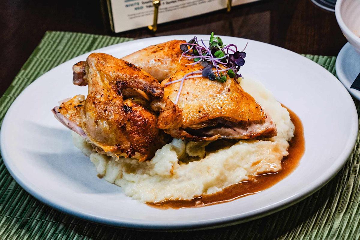 Roast chicken with crispy skin served on a bed of mashed potatoes, topped with microgreens, and surrounded by brown gravy on a white plate.