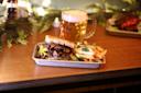 A tray on a wooden table holds a sandwich filled with slices of beef, lettuce, and pickles, accompanied by a side of French fries. A mug of frothy beer is in the background, with decorative greenery nearby.