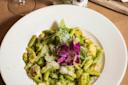 A white plate of pasta with a creamy green pesto sauce, grated cheese, and chunks of chicken. A purple flower garnishes the dish, adding a pop of color. A drink and some condiments are blurred in the background.