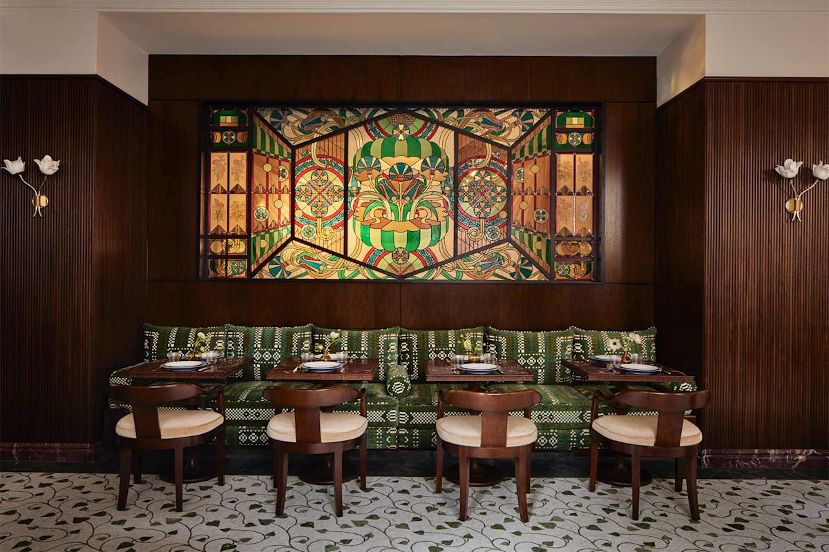A dining area with dark wood walls, patterned green bench seating, four wooden chairs, set tables, and a large colorful geometric mural above the seating. The floor has a light tile with a floral pattern.