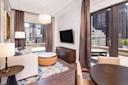Modern living room with large windows and city views. Features a beige sofa, round coffee table, flat-screen TV on the wall, and a round dining table. Elegant pendant light hangs from the ceiling. Dark curtains and a patterned rug complete the look.
