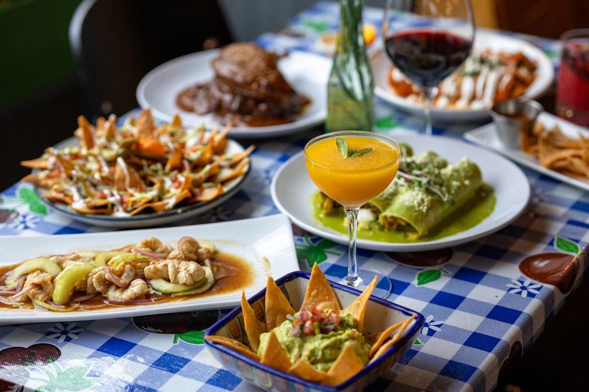 A colorful table set with various Mexican dishes, including guacamole with chips, enchiladas, nachos, seafood, a yellow cocktail, and a glass of red wine, all on a blue checkered tablecloth.