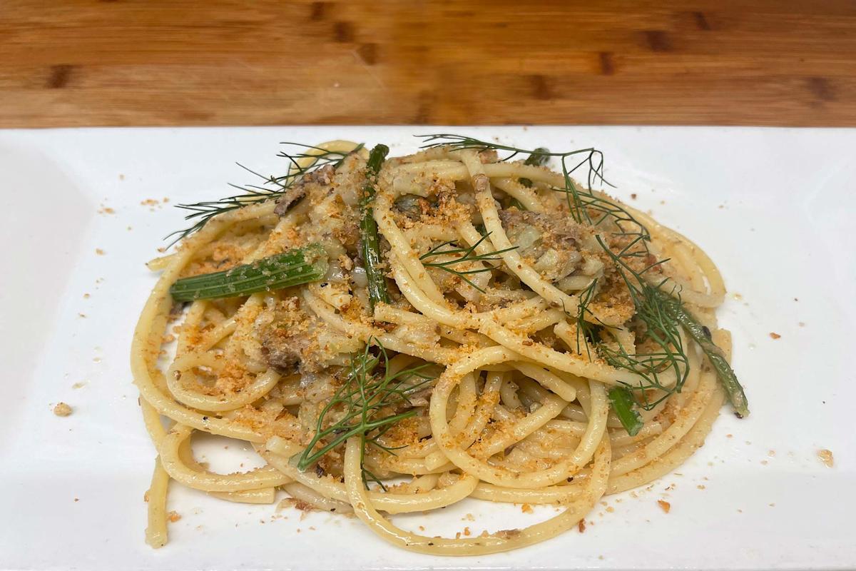 A plate of pasta with sausage, green beans, and herbs, garnished with fresh dill and breadcrumbs, served on a white rectangular dish on a wooden table.