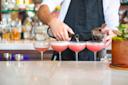A bartender in a white shirt and black apron pours liquid from a bottle into one of four pink cocktails lined up on a marble bar counter. The background features shelves with various bottles and cocktail shakers.