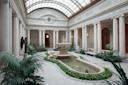 Elegant indoor courtyard with a central stone fountain, surrounded by lush plants, white columns, statues, and a glass-domed ceiling allowing natural light to fill the classical architectural space.