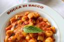 A plate of gnocchi in tomato sauce garnished with a fresh basil leaf, served on a white plate with "Bocca di Bacco" written around the rim. The dish is placed on a white tablecloth.