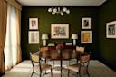A sophisticated dining room with a round wooden table, four chairs, flower centerpiece, dark green textured walls, modern chandelier, framed abstract art, sideboard with lamps, books, and floor-to-ceiling gold curtains.