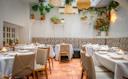 A bright, elegant restaurant dining area with white tablecloths, neutral chairs, potted plants, hanging woven lights, and patterned bench seating against a white brick wall. Candles are lit on each table.
