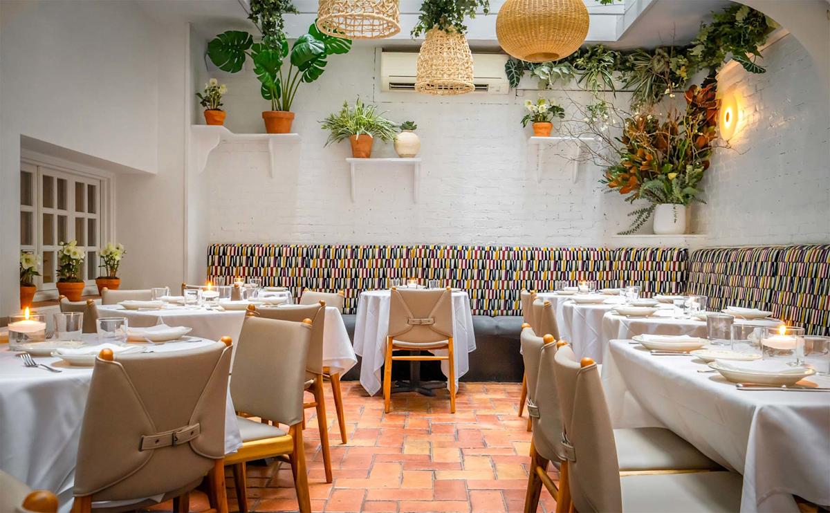 A bright, elegant restaurant dining area with white tablecloths, neutral chairs, potted plants, hanging woven lights, and patterned bench seating against a white brick wall. Candles are lit on each table.