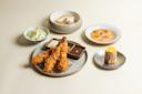 A meal featuring fried shrimp with dipping sauces, a bowl of sliced cabbage, grilled fish in broth, thinly sliced fish carpaccio with orange, and a piece of grilled corn on a small plate, all on a light background.