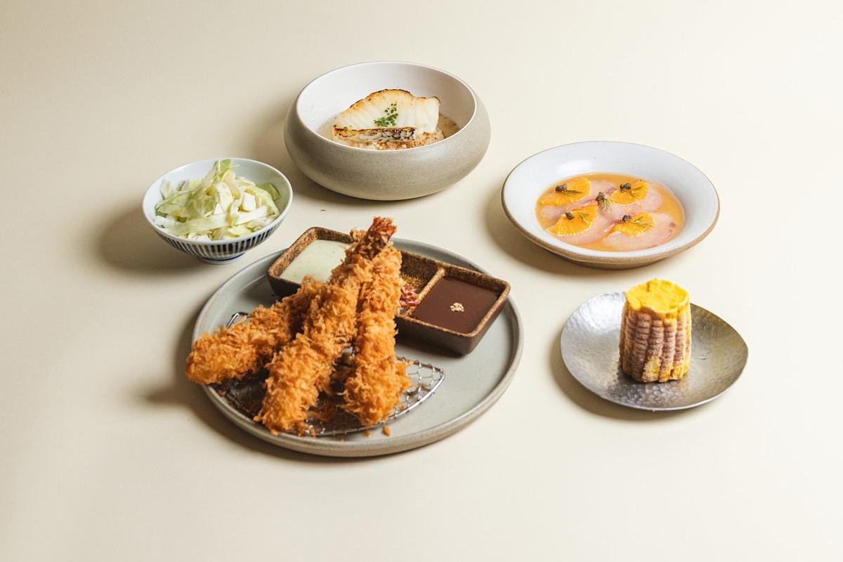 A meal featuring fried shrimp with dipping sauces, a bowl of sliced cabbage, grilled fish in broth, thinly sliced fish carpaccio with orange, and a piece of grilled corn on a small plate, all on a light background.